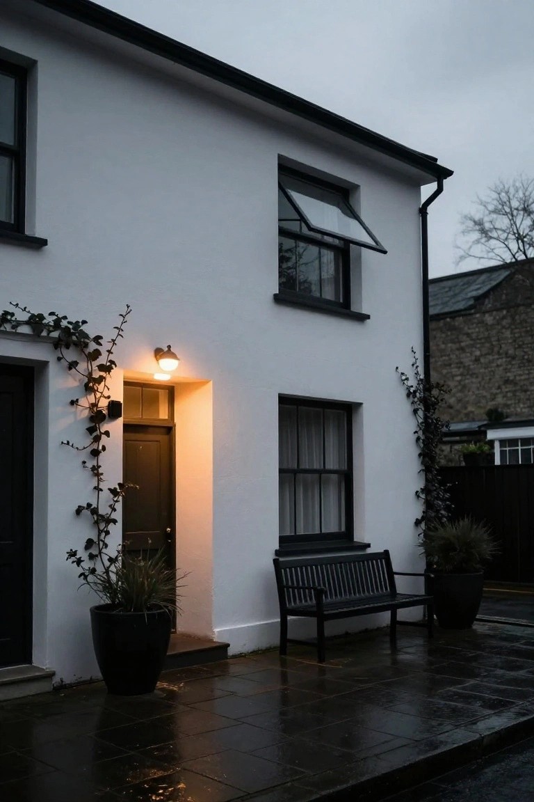 White stucco house exterior corner featuring a black front door illuminated by a black wall-mounted light, ivy on the wall, potted grasses, black bench, and wet slate paving at dusk.