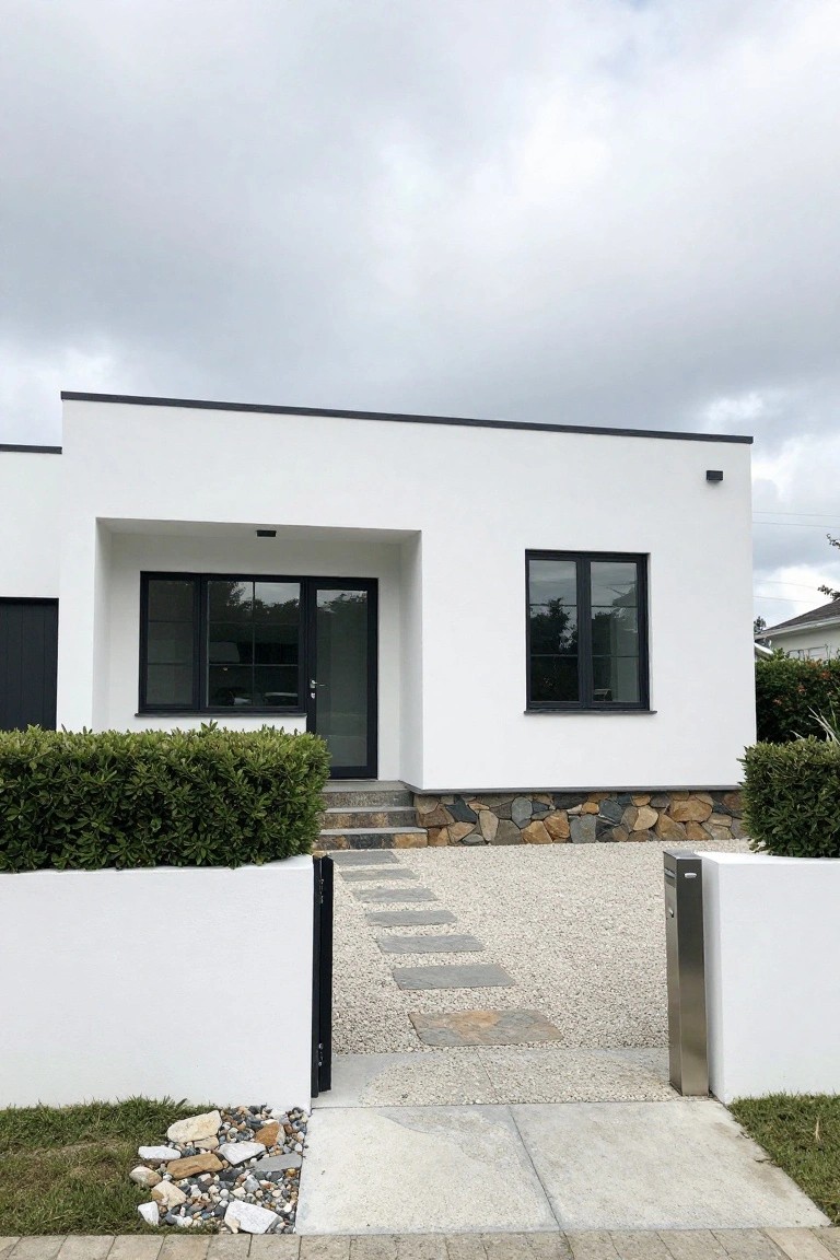 Modern single-story white house with black-framed windows and door, low boxwood hedges along white boundary walls, gravel area with stepping stone path leading to entry, stone accents at base under cloudy sky.