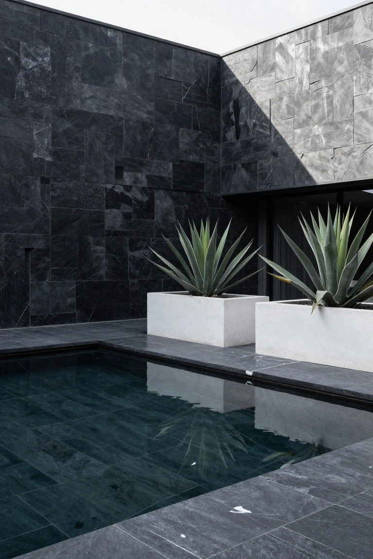 Corner view of modern home exterior with dark slate walls, small dark green pool, and two large white square planters containing agave plants.