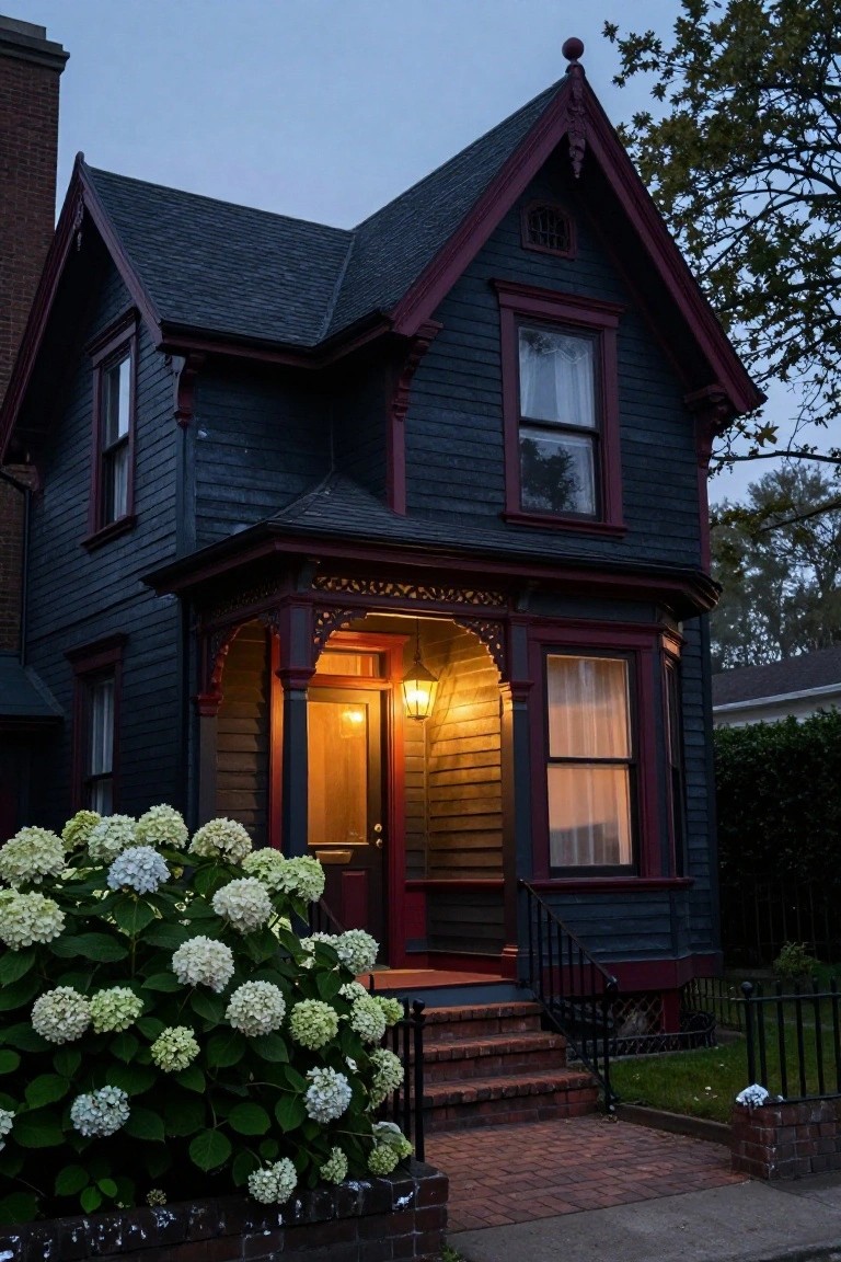 Dark shingled two-story Victorian house with red trim and an ornate wooden front porch featuring a lit lantern, flanked by large white hydrangea bushes and brick steps, at dusk.