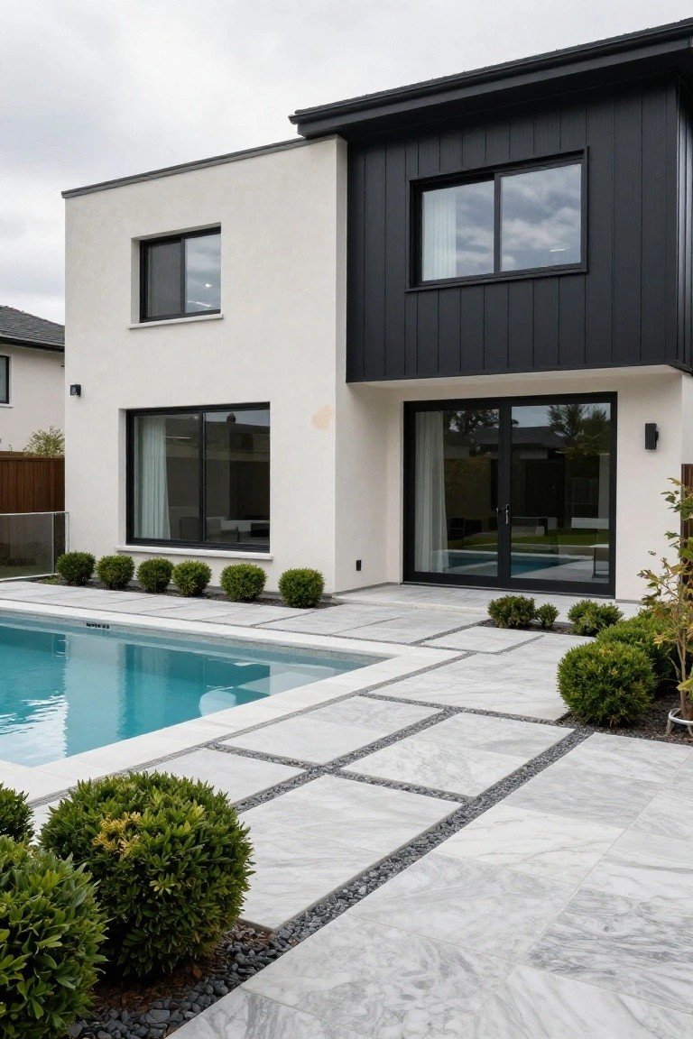 Modern house exterior with white and black walls, large black-framed sliding doors opening to a rectangular pool, and an adjacent patio paved in large white tiles separated by black pebble lines, edged with boxwood shrubs.
