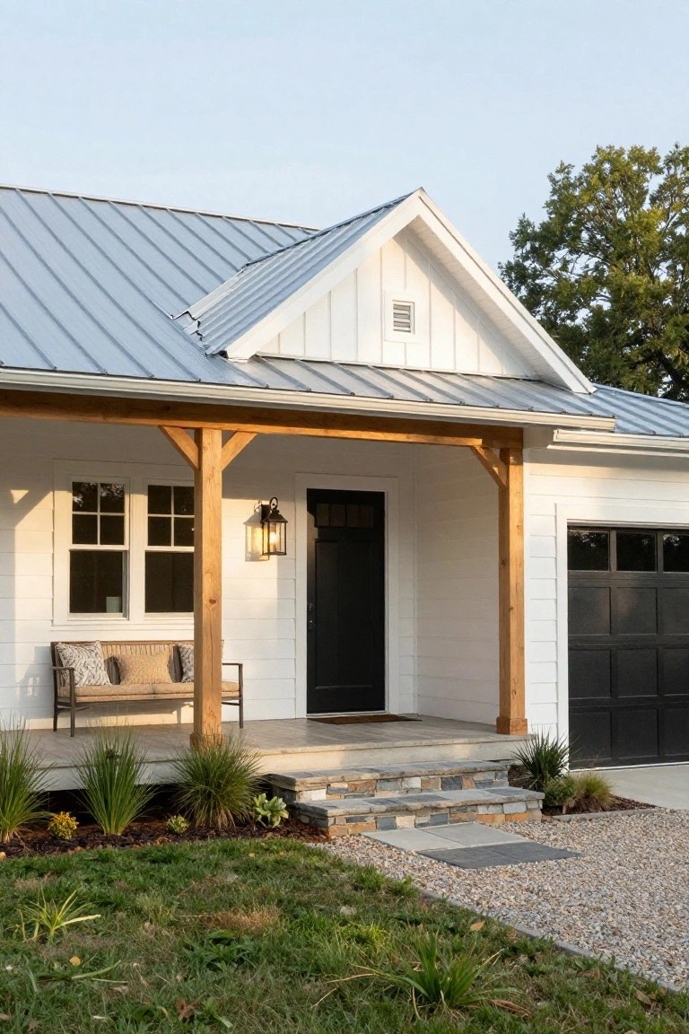 White modern farmhouse with gray standing seam metal roof, wooden porch beams and posts, black front door and garage door, porch bench, stone steps to gravel path, and grass with plants in front yard.