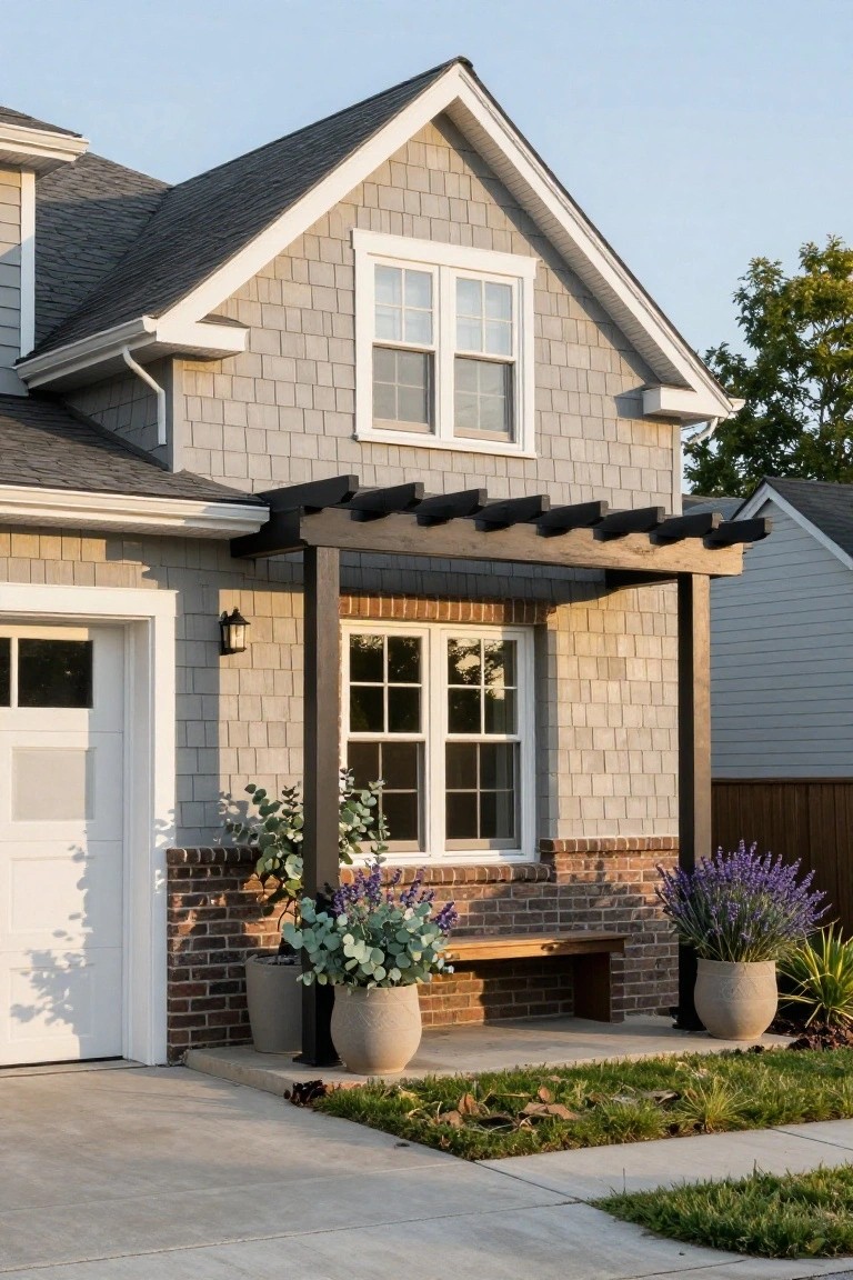 Gray shingle siding house with attached white garage door, black wooden pergola over brick bench and large potted eucalyptus and lavender plants next to concrete driveway.