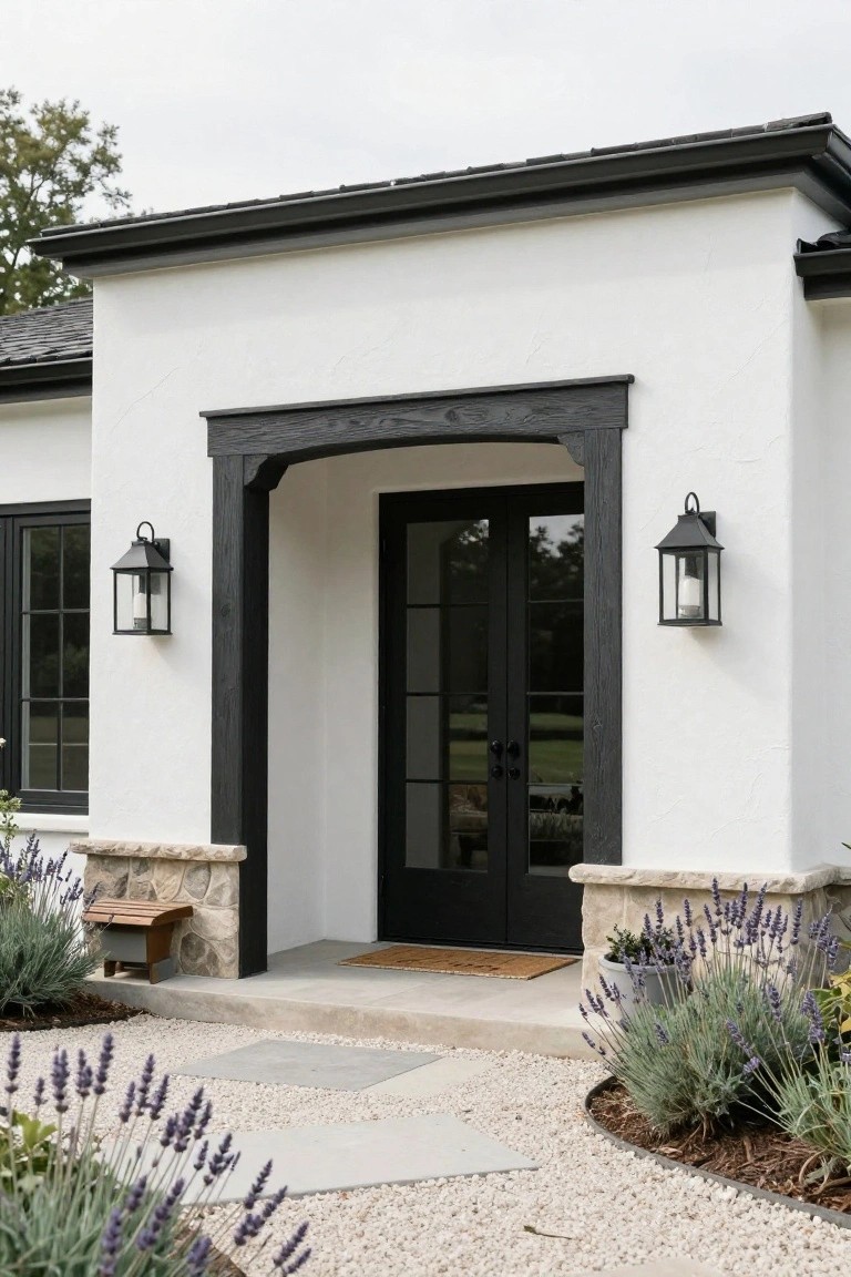 A white stucco house exterior featuring a dark wood arched entry framing black double doors, with black lanterns on either side and stone bases holding lavender plants.