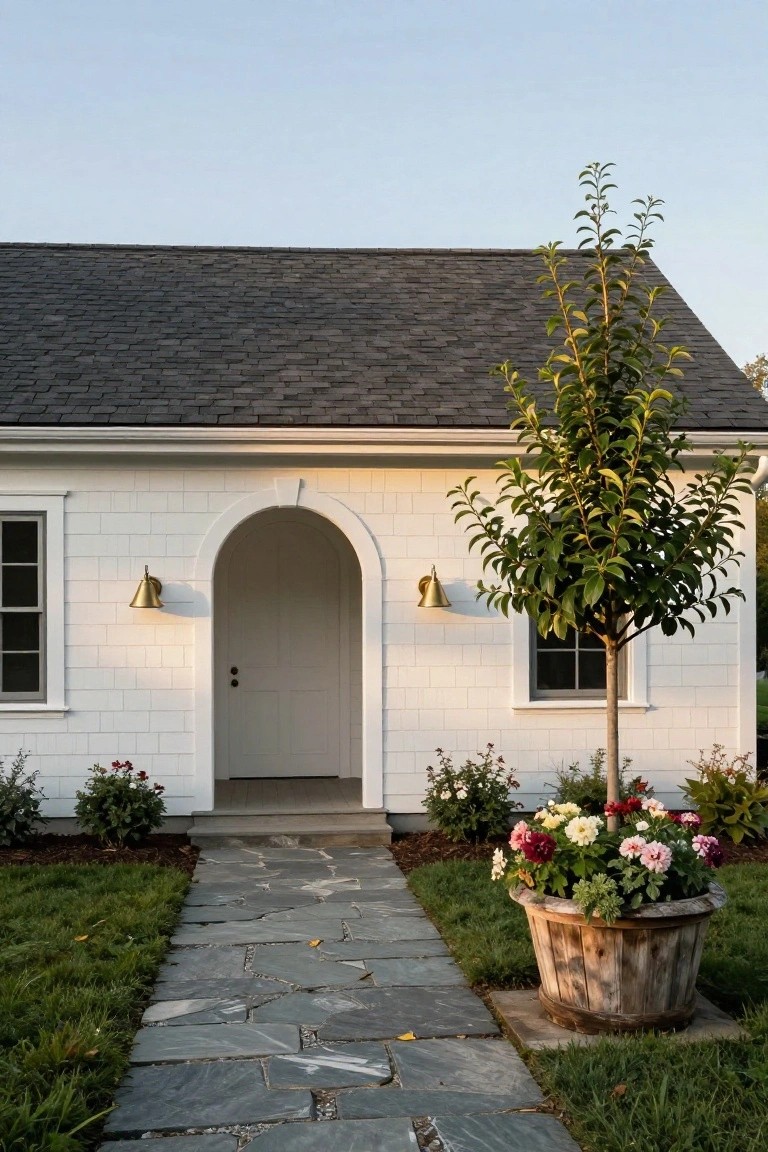 A white one-story house with dark shingles has an arched front door, two brass wall sconces, a stone path, and a wooden planter with flowers to the right.