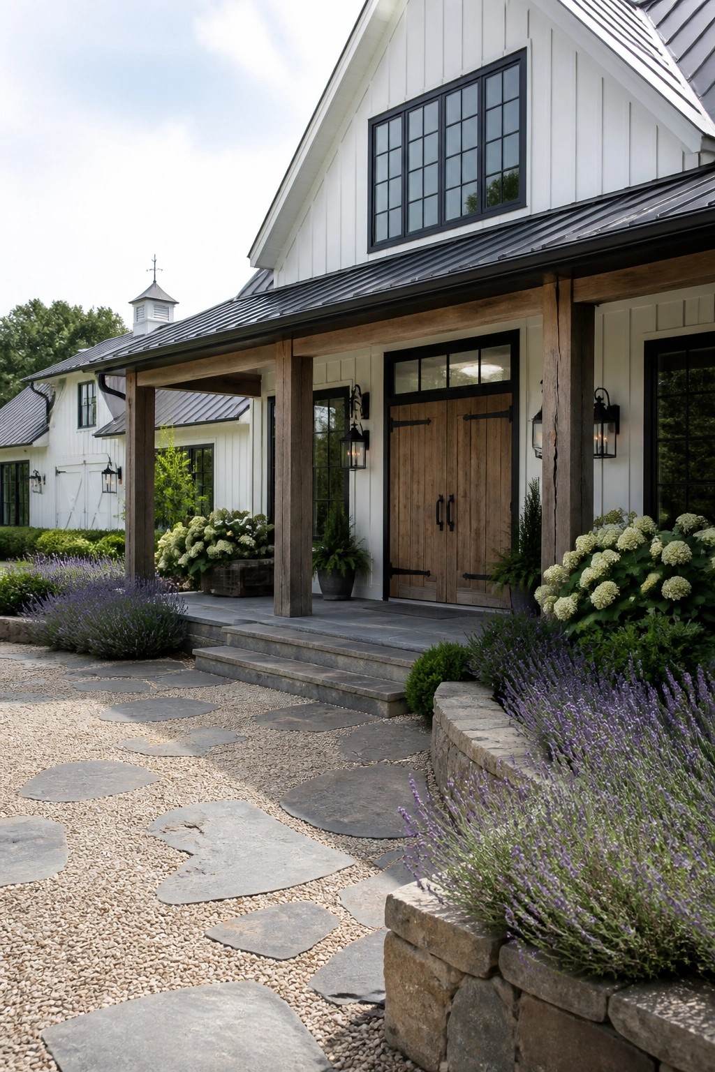 A white house exterior with a wooden porch supported by thick posts, double wooden front doors, black-framed windows, and a stone pathway bordered by lavender.