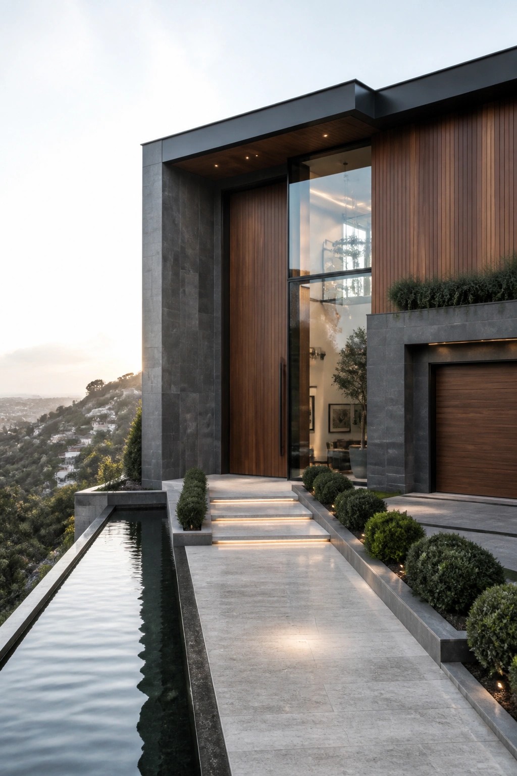 A modern house exterior with a long rectangular water feature running parallel to a paved walkway that leads to a tall wooden front door set in stone walls.