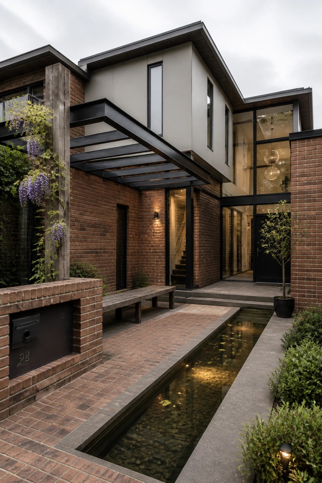 Modern two-story house exterior with brick walls, a black metal canopy extending over the entry path, and a narrow water feature running alongside the brick walkway.