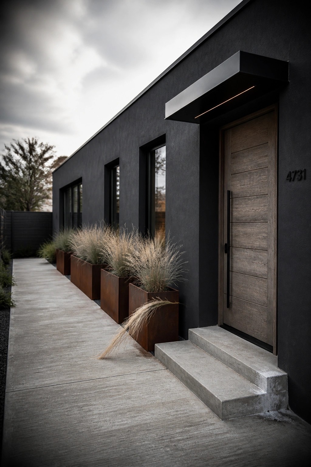 A modern one-story house exterior with dark gray walls, a vertical wood front door, concrete steps, and a row of tall grasses in rusted metal planters along the walkway.