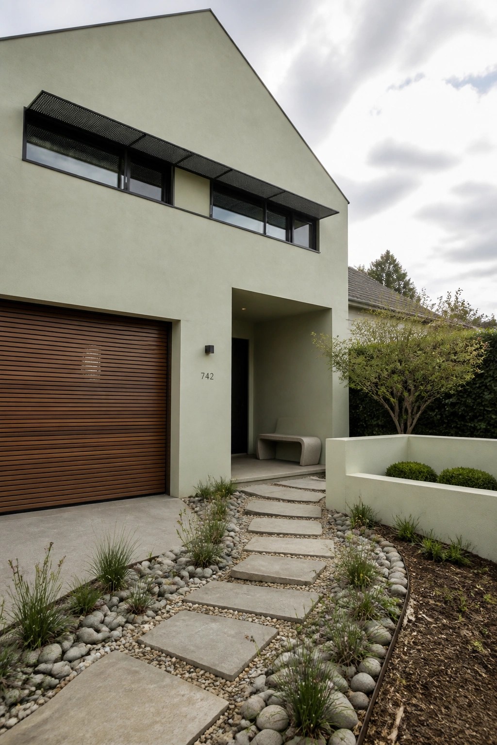 Modern two-story house with light green stucco exterior, brown slatted garage door on the left, recessed entry with dark door and concrete bench on the right, and a stone paver path through gravel landscaping.