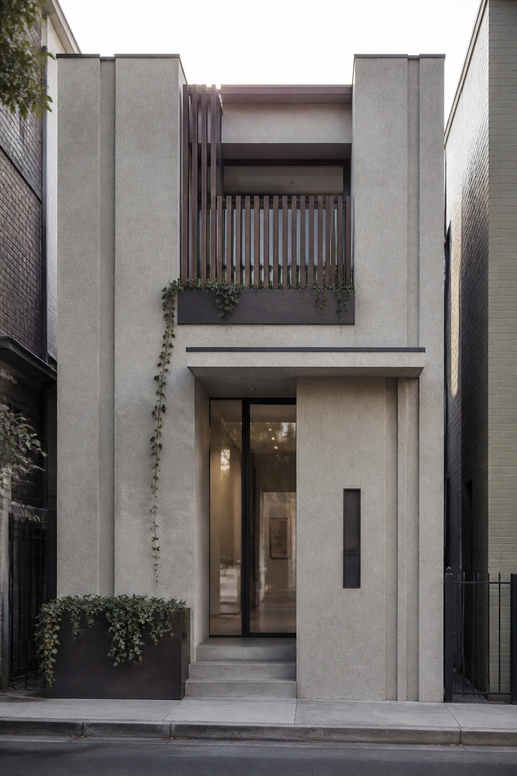 Front view of a narrow modern concrete house with a recessed glass entry door and a balcony fitted with vertical wooden slats above it.