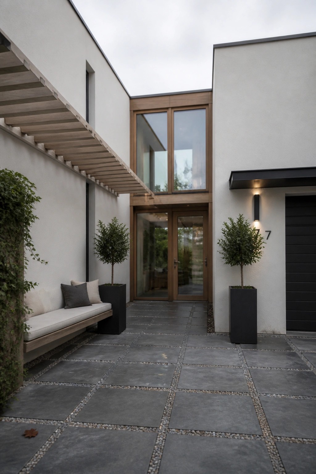 A minimalist courtyard with gray stone pavers, a built-in wooden bench with cushions, and two tall black planters holding small trees beside modern glass doors.