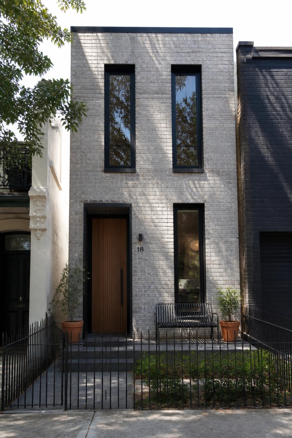 A narrow gray brick house with tall black-framed vertical windows, a wooden front door, and a small fenced entry area.
