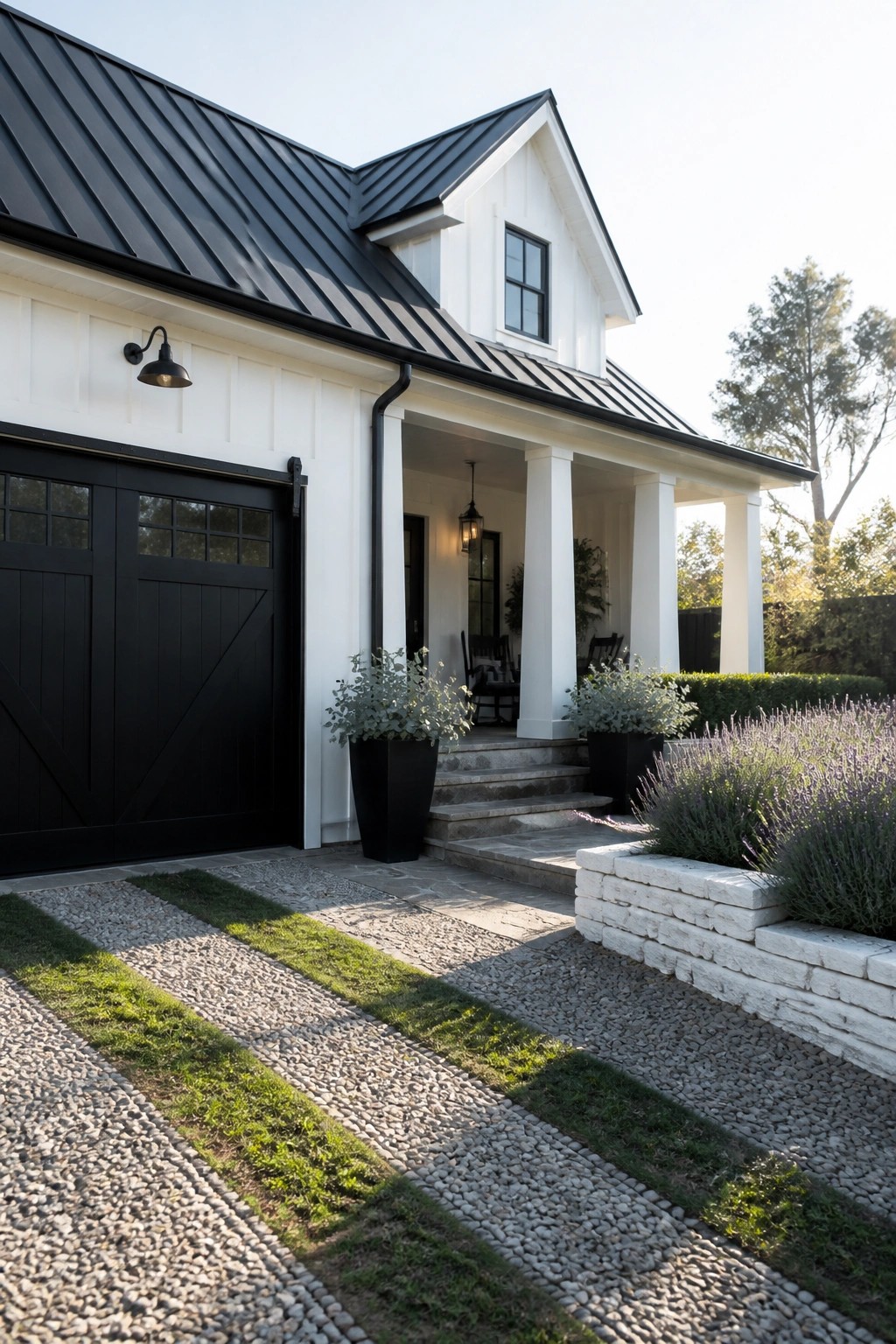 A modern farmhouse exterior with white siding, a black metal roof, black garage door, white porch columns, stone steps, and gravel landscaping with potted plants.