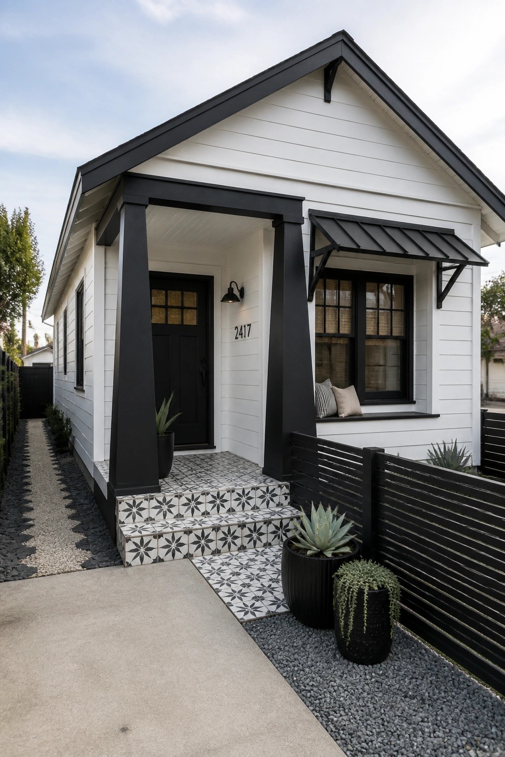 A small white house with black trim around the porch, door, and windows, featuring tiled steps and potted plants at the entrance.