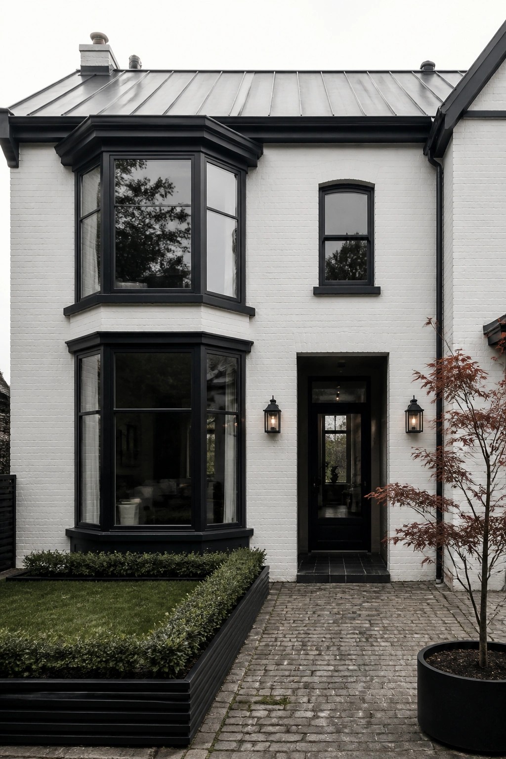 A white brick house exterior featuring prominent black-framed bay windows, a black front door, and a cobblestone walkway with black planters.