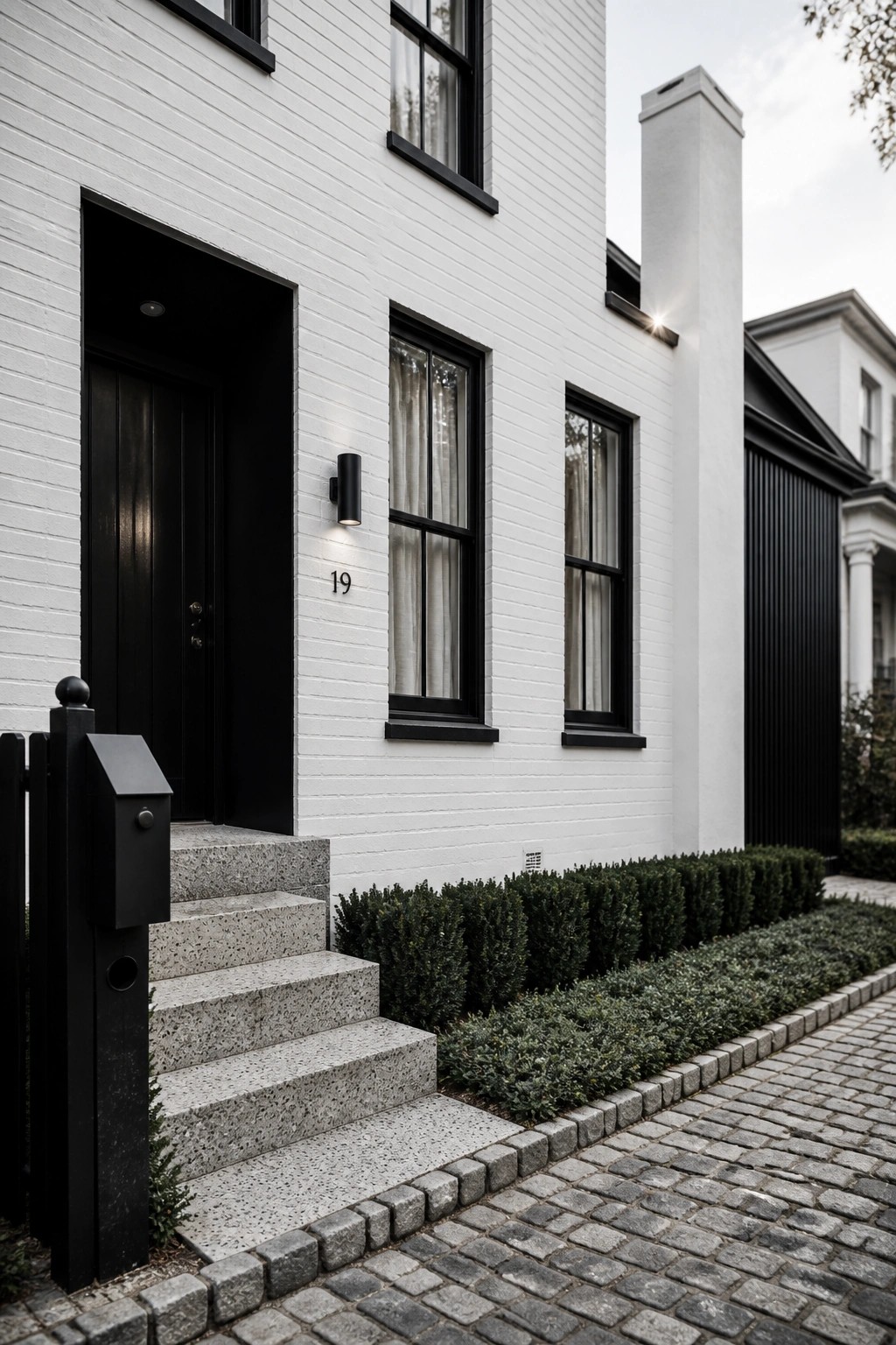 Front view of a white brick house with black-framed windows, a black door, stone steps, and a black mailbox post beside a cobblestone path.
