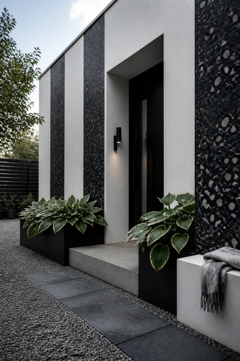 A modern house entry with black and white vertically striped walls, a black door, concrete steps, and large leafy plants in black planters.