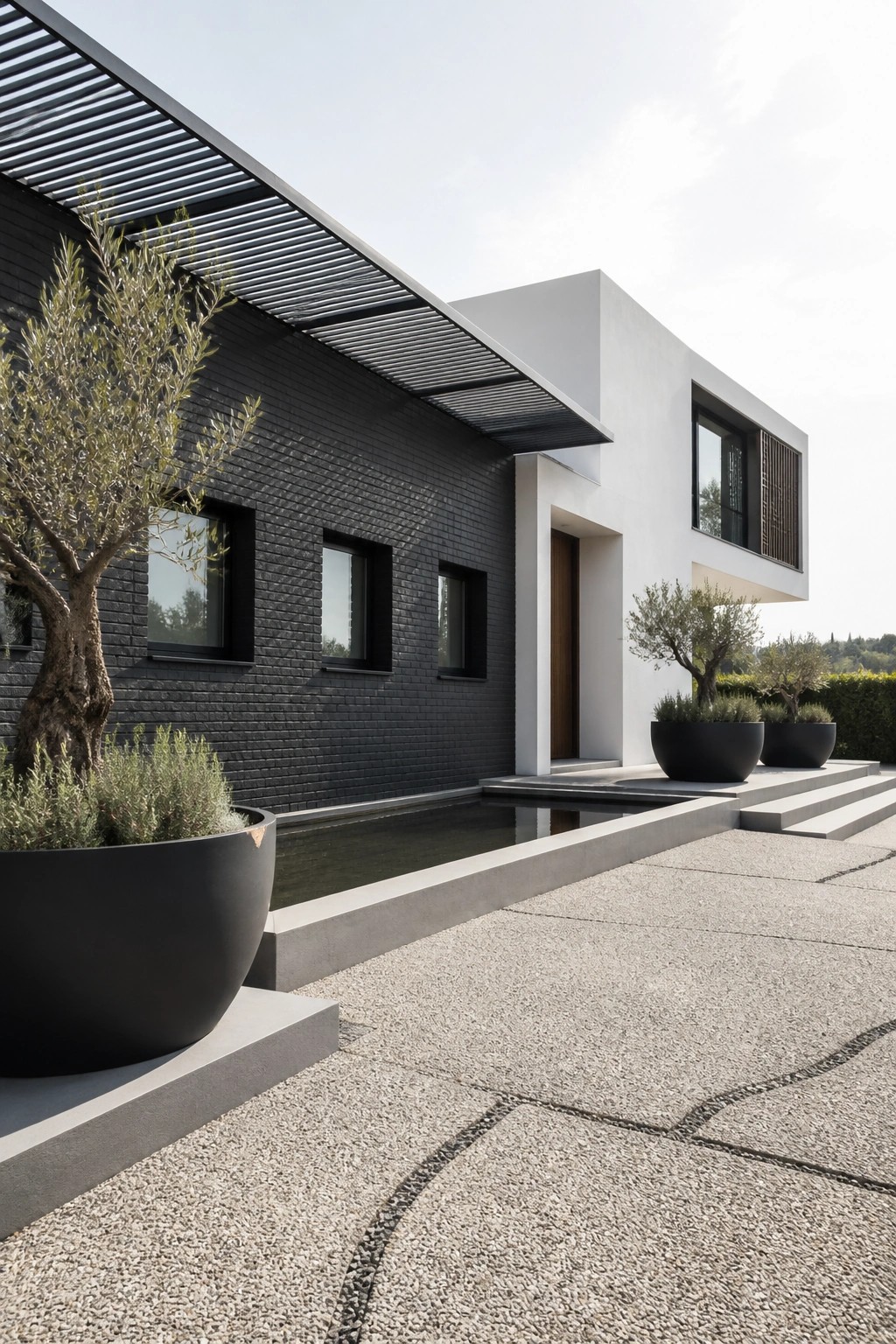 A modern house exterior with a dark brick facade featuring small rectangular windows, adjacent white geometric volumes, a long narrow pool, and black planters holding trees on a gravel area.