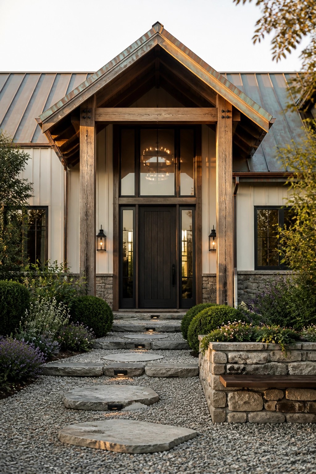 A modern farmhouse exterior with a steep gabled entry supported by heavy timber posts, a dark front door with glass sidelights, stone foundation, and a gravel path with stepping stones.