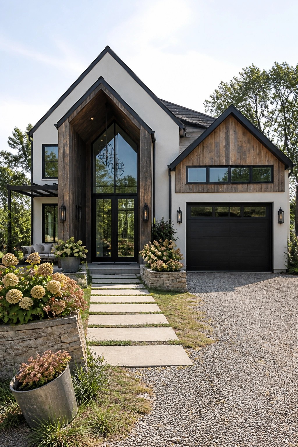 A modern farmhouse with white siding, a tall dark wood gabled entry framing large glass doors, black trim, and a gravel driveway bordered by stone planters.