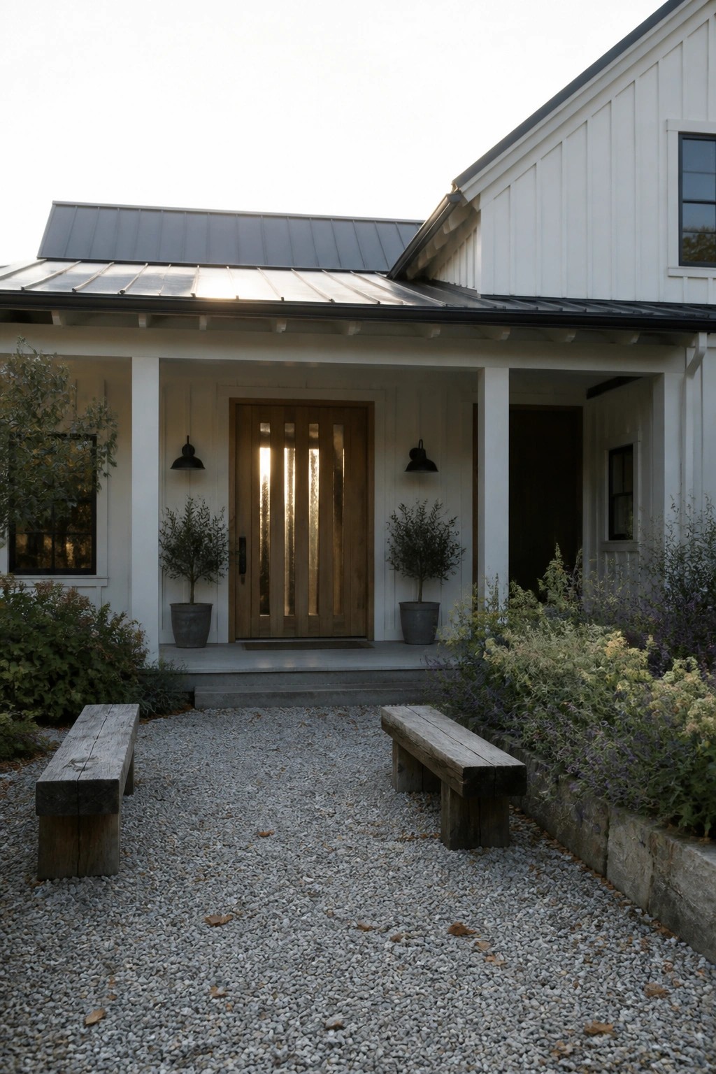 White house exterior with a wooden front door featuring vertical glass panels, two potted shrubs on either side, and a gravel area with wooden benches in front.