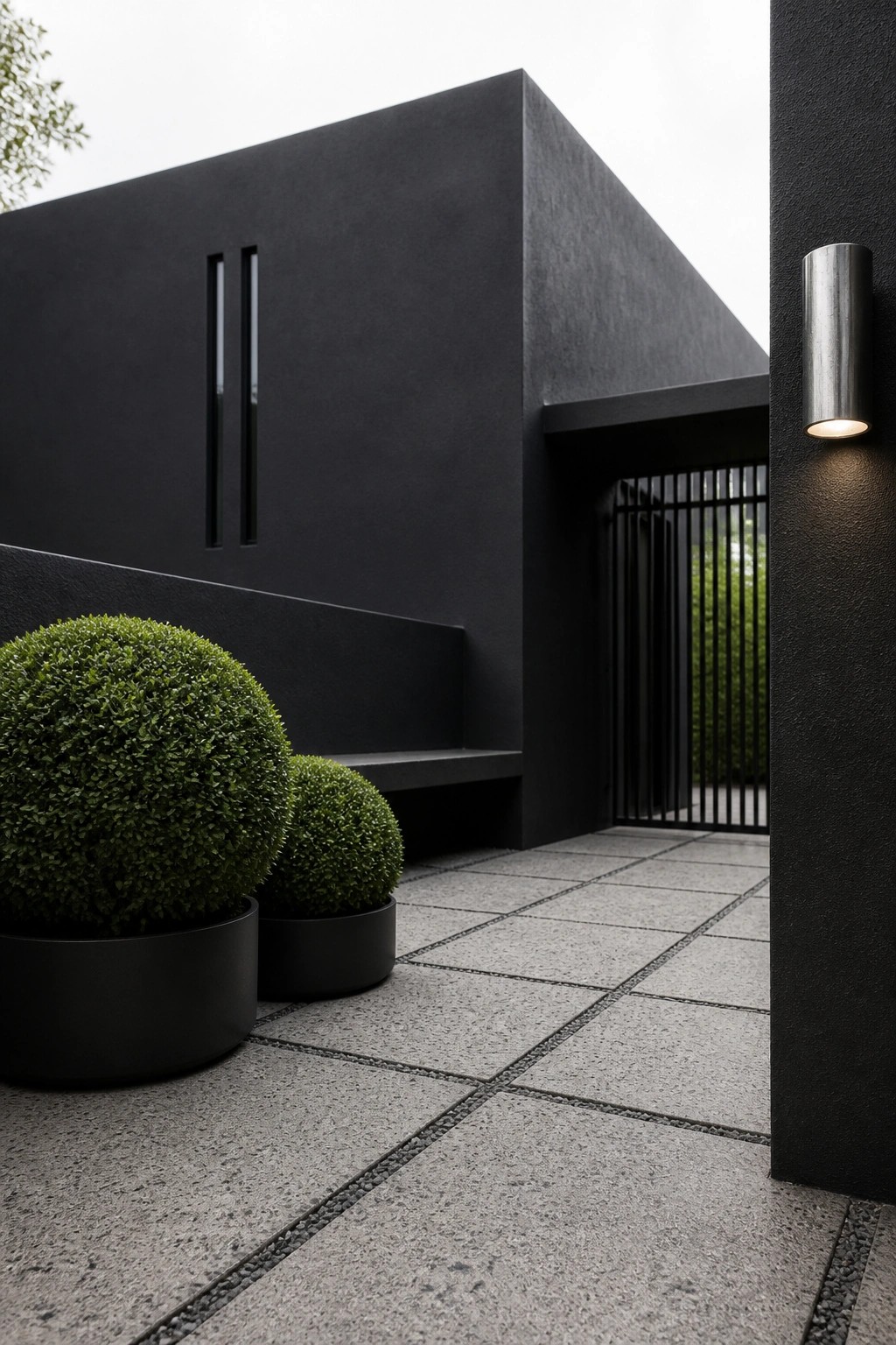Modern black building with stone tiles, potted shrubs, bench, and wall sconce.