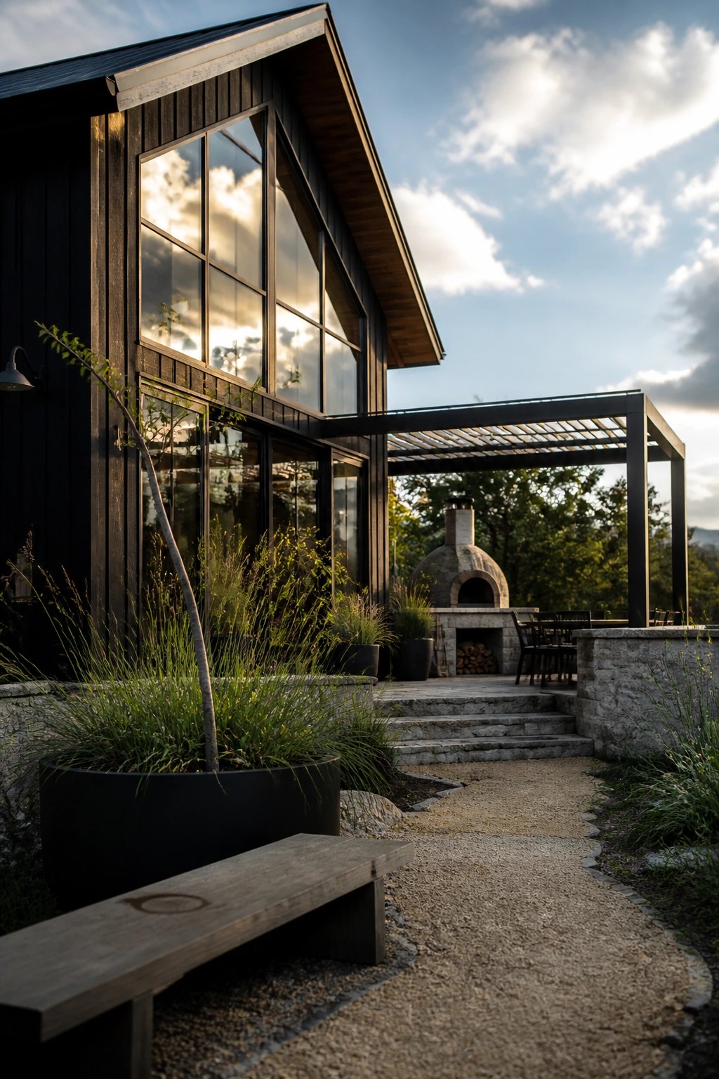 Modern black house with vertical siding and large multi-pane windows next to a covered patio with stone oven and pergola.