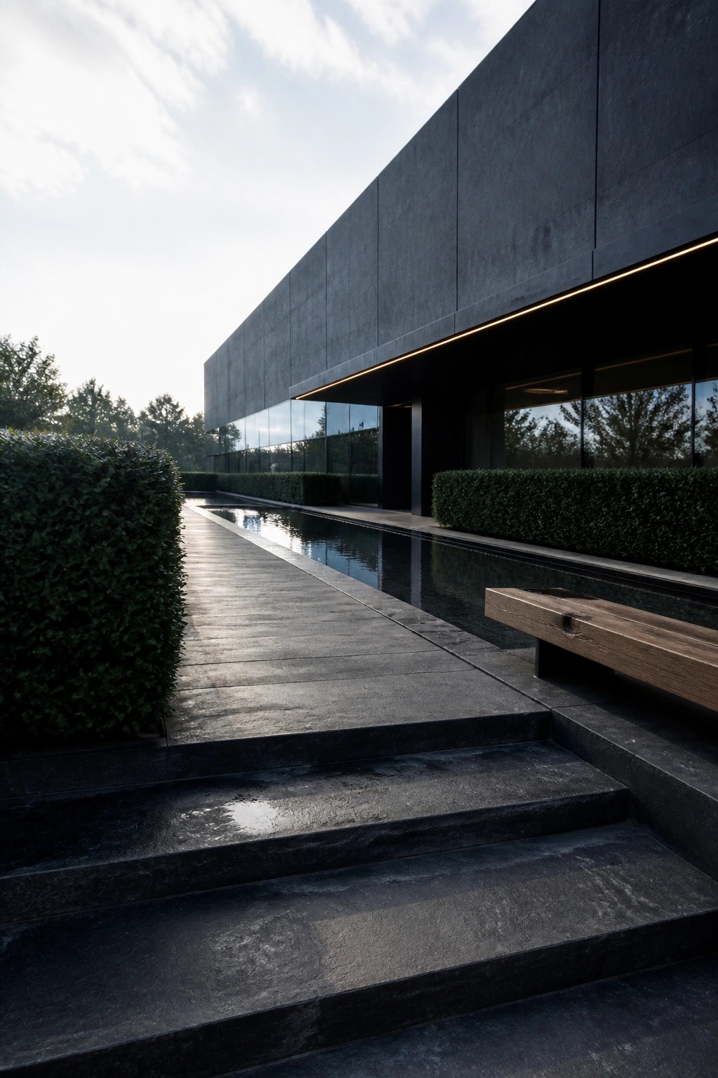 Dark concrete building with long reflecting pool, stone steps, wooden bench and hedges