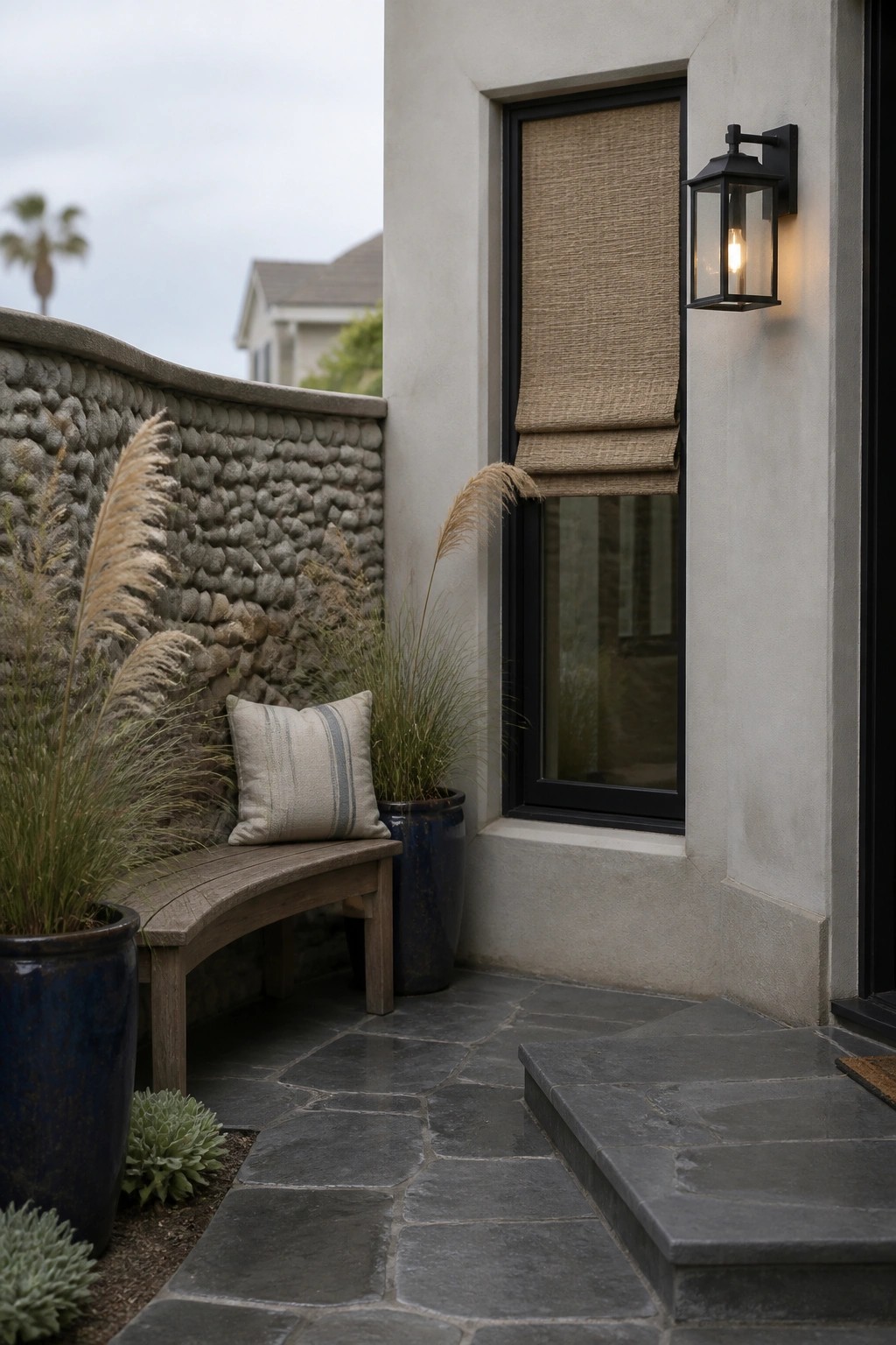 A curved wooden bench with a pillow sits against a stone wall beside a window with a rolled shade on a paved outdoor area.