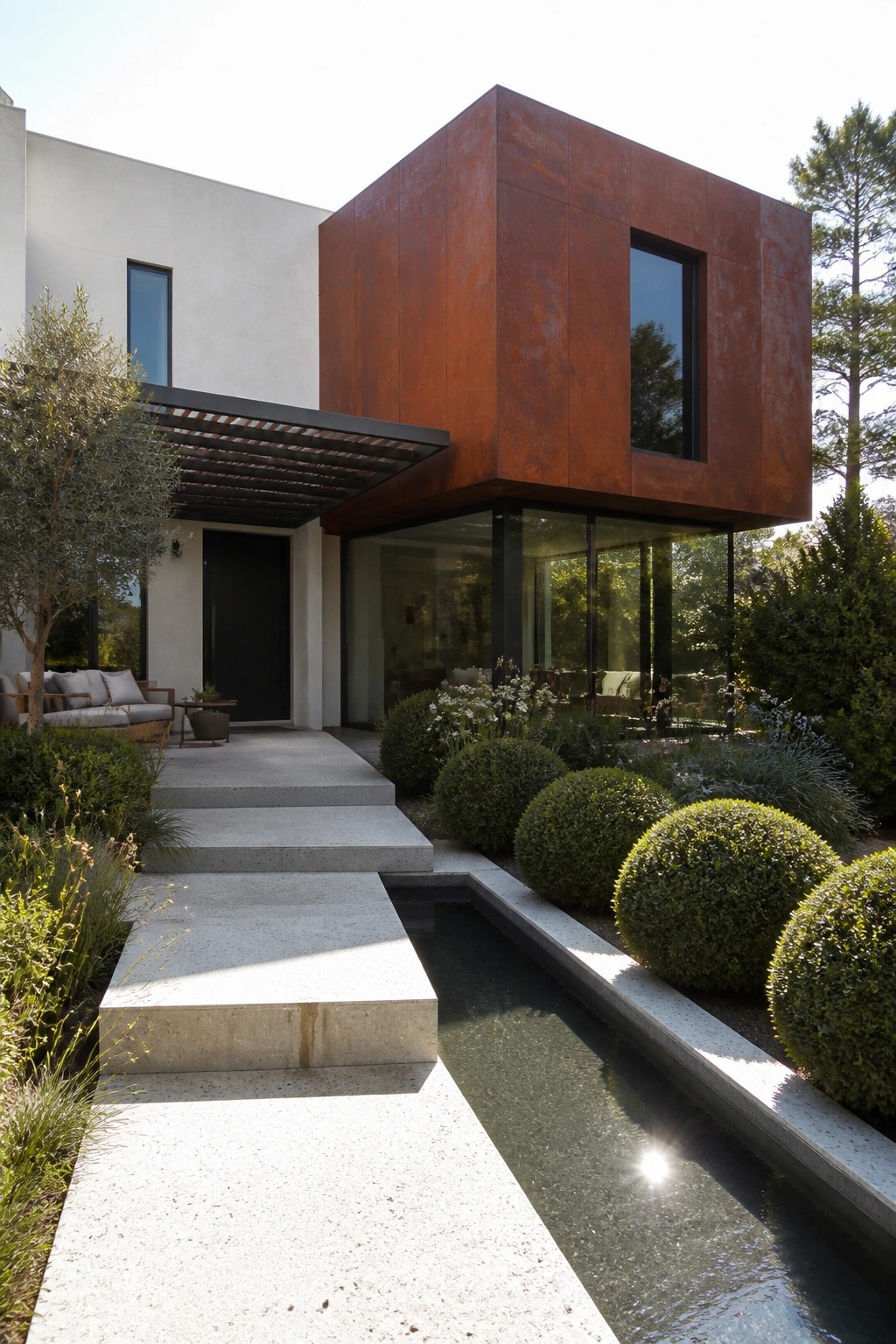 A modern house exterior with a large rusted metal cube on top of a white stucco base, glass walls below, concrete steps, and a narrow water channel along the walkway.