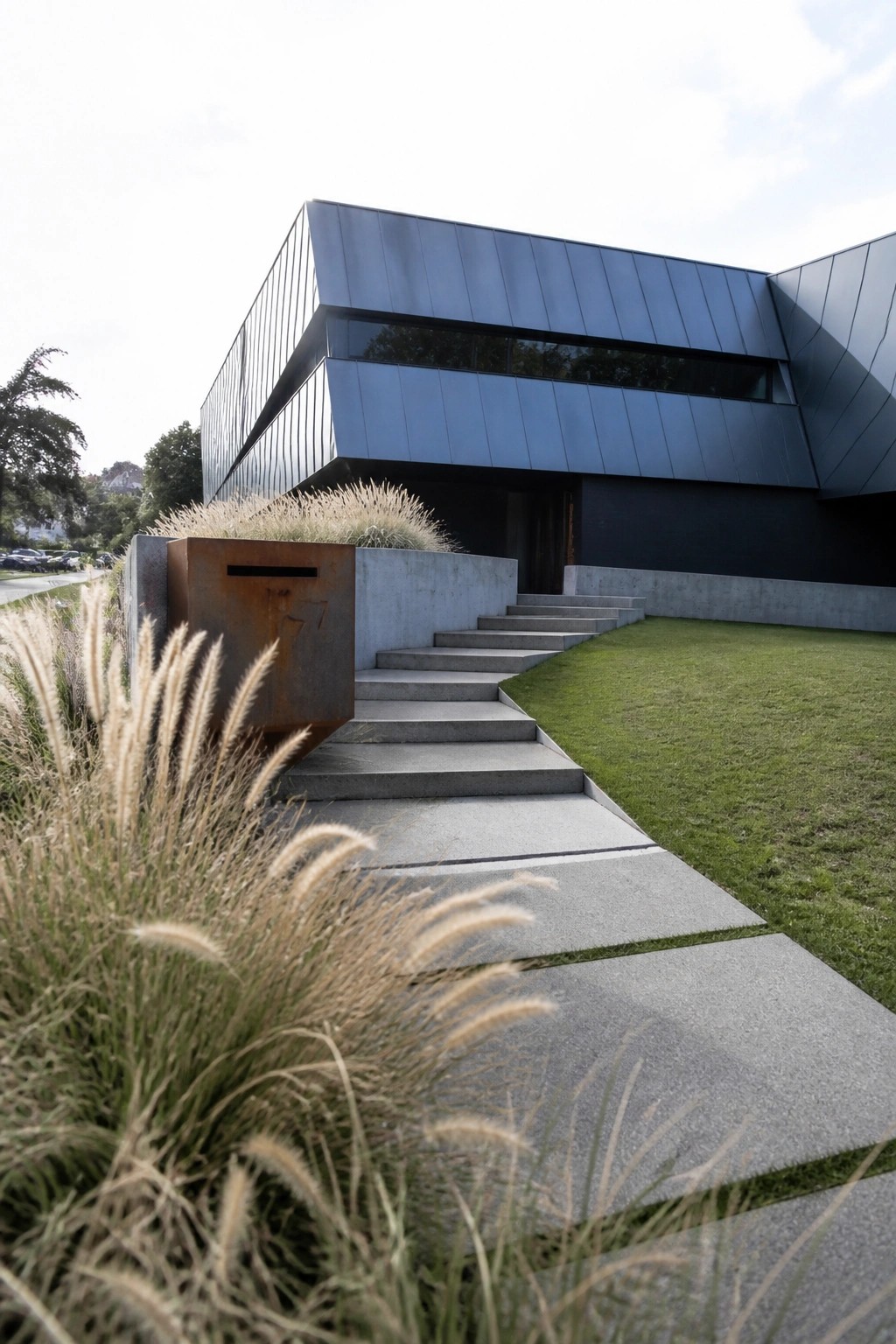 A contemporary house with angular blue-gray metal cladding, concrete entry stairs, and tall ornamental grasses beside a rusted metal mailbox structure.