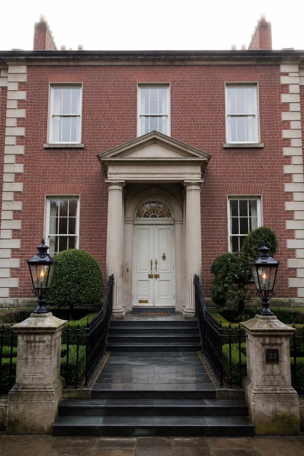 A red brick house with a central white double door framed by two stone columns and a triangular pediment, accessed by wide stone steps with black railings and lanterns on either side.