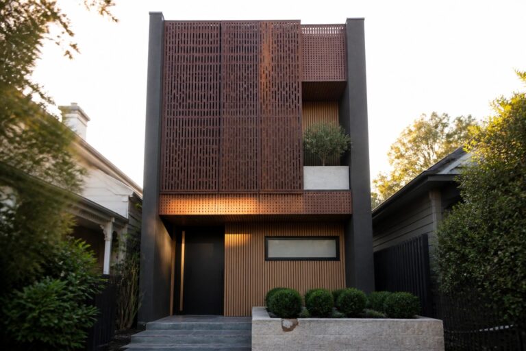 Modern narrow house with dark walls, a large vertical wooden slat screen on the upper facade, and a covered entry with steps.
