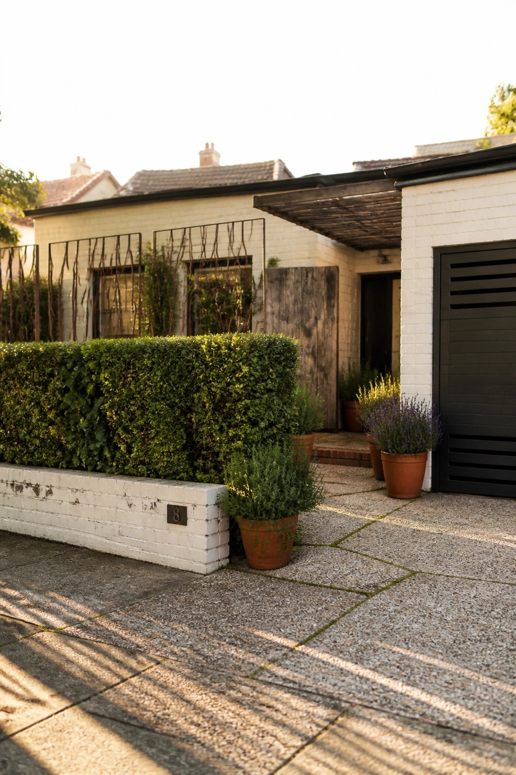 A white brick house front with a tall dense green hedge along the boundary, terracotta pots with plants, and a black door under a wooden pergola.