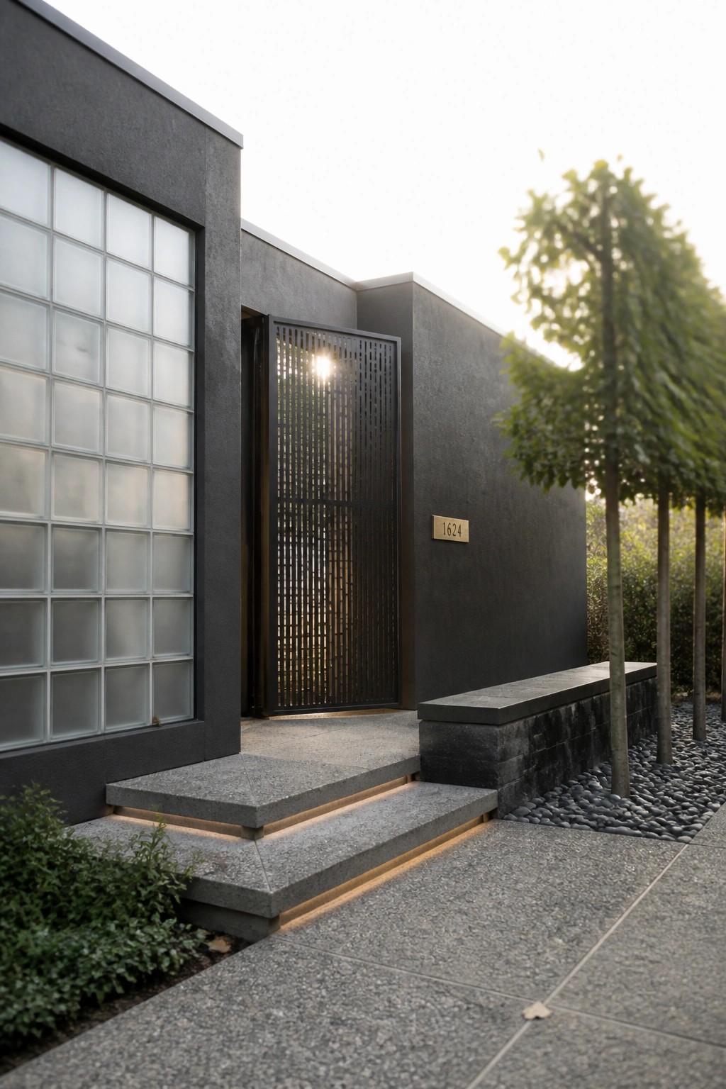 A modern dark house facade with a vertical slatted metal gate at the entrance, a glass block window on the left, stone steps with lighting, and a gravel bed with trees to the right.