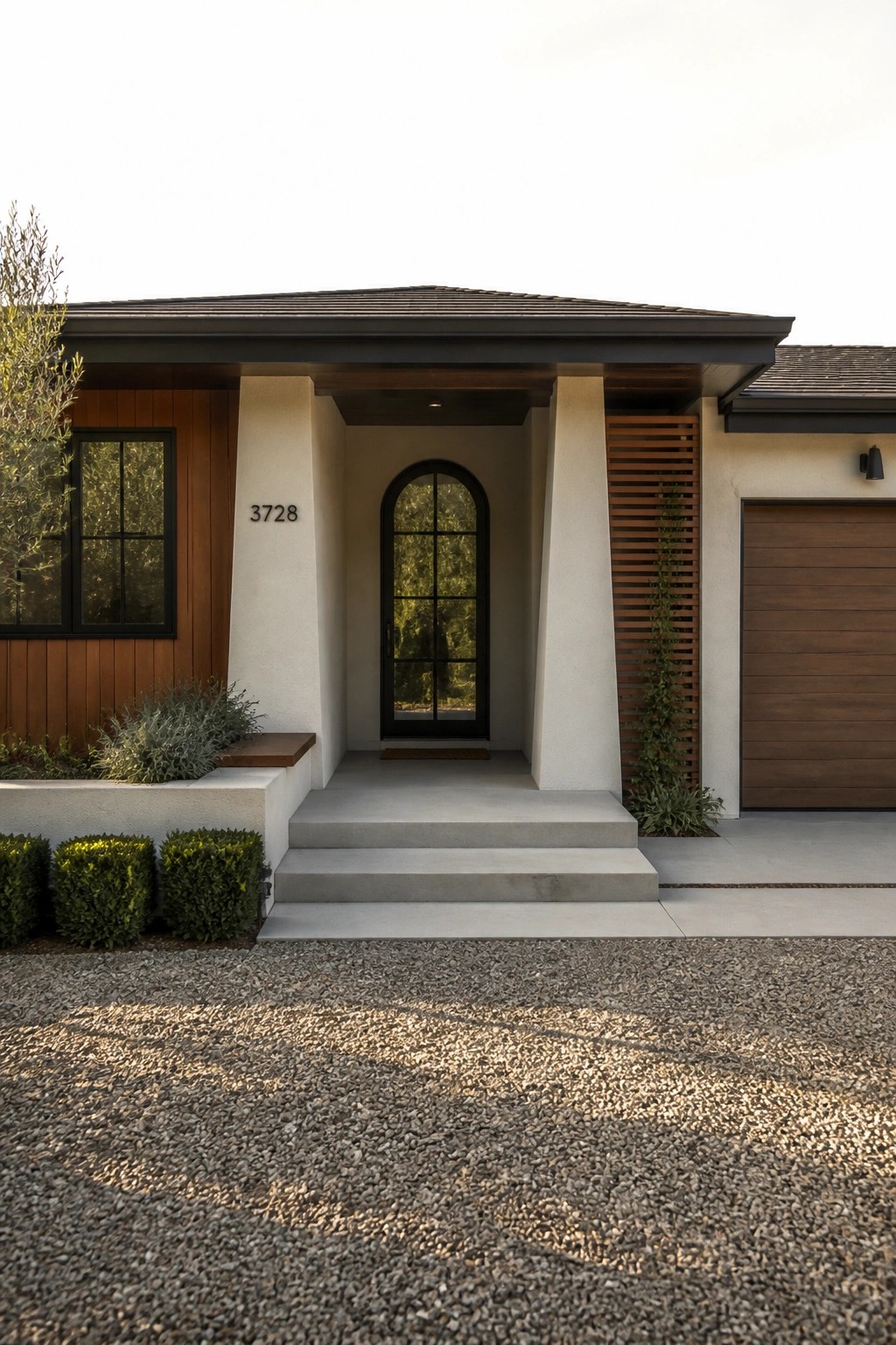 Front view of a modern bungalow with a covered porch supported by white columns, an arched black door, wood siding on the left side, and a garage door on the right.