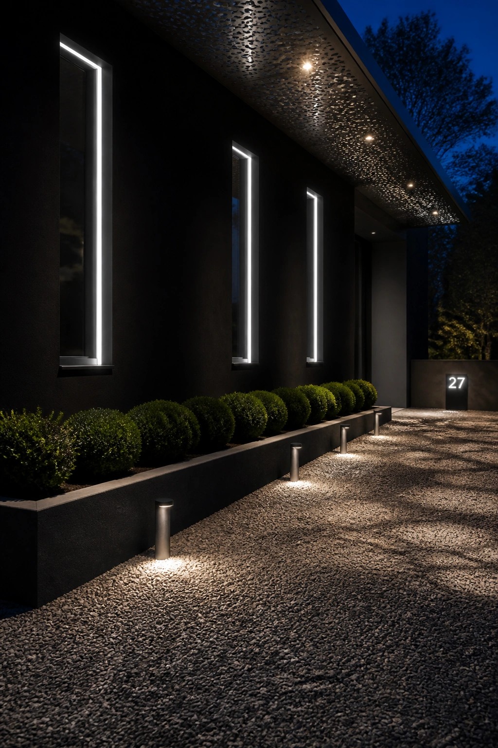 Modern dark house exterior at night showing three tall windows framed by vertical LED light strips, with bollard lights along a gravel path and low shrubs.