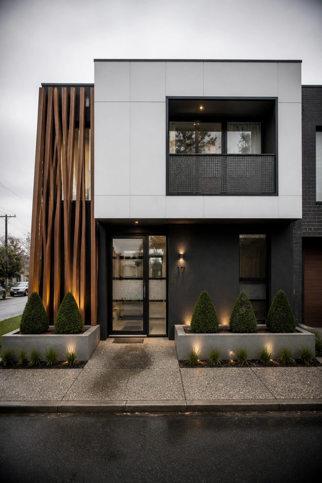 A modern two-story flat roof house with white upper panels, a dark lower section, vertical wood slats on the left side, black-framed glass doors, and concrete planters with shrubs along the front.