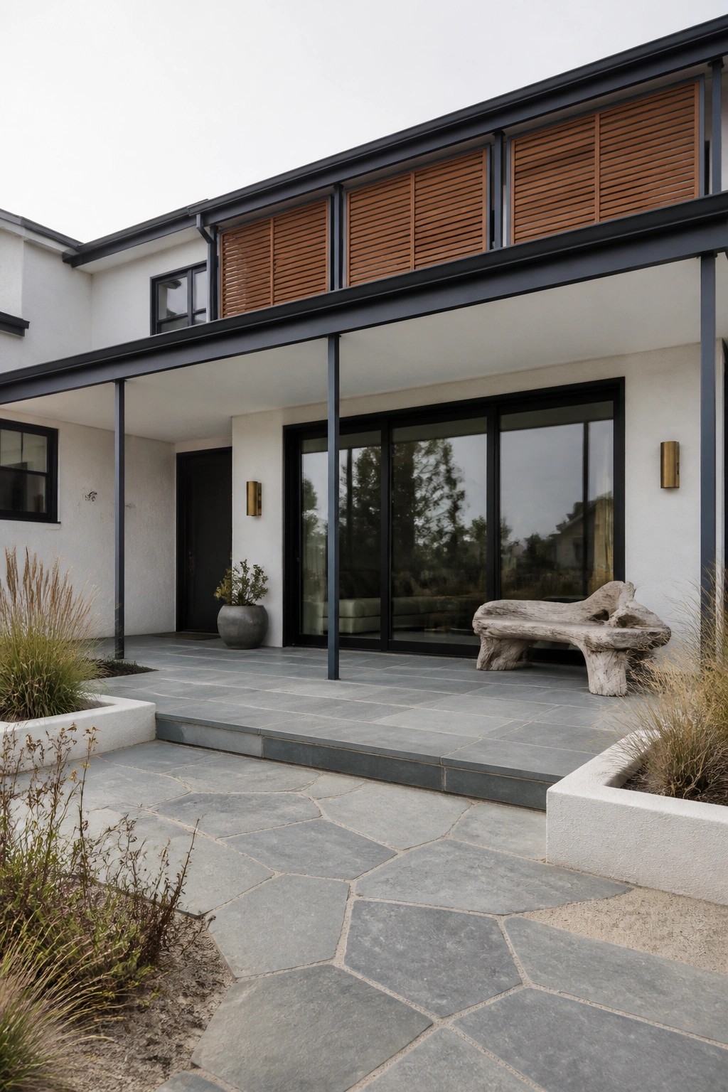 Modern white house exterior with black metal trim, large sliding glass doors, covered patio, and horizontal wood shutters on the upper level above a stone paver walkway.