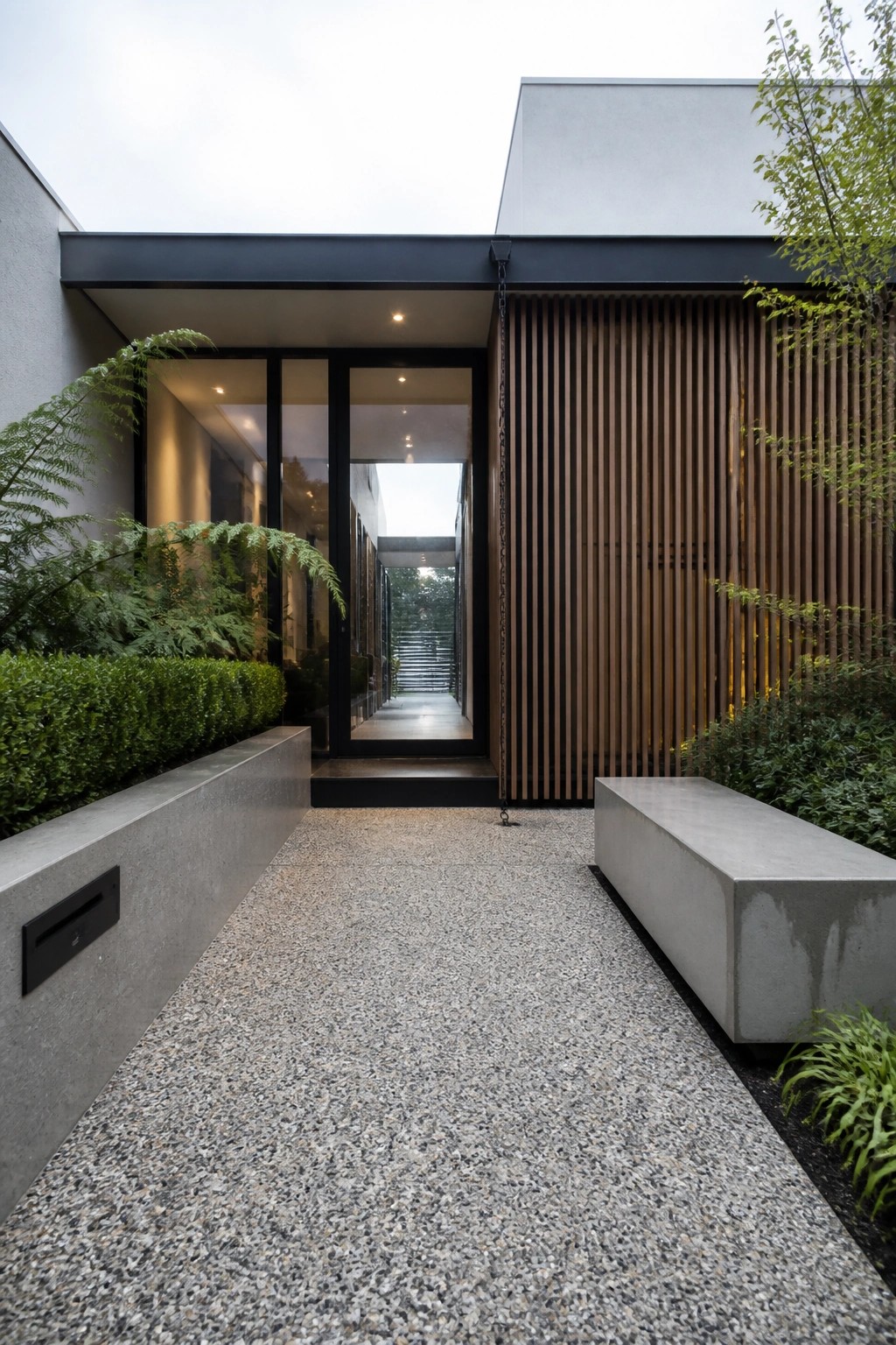 Modern minimalist entrance with gravel path, glass doors, concrete benches and wood slats.