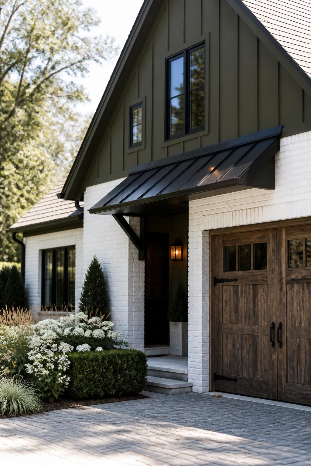 A front view of a house showing dark green vertical siding, white brick walls, wooden garage doors, and a black metal awning above the entry.