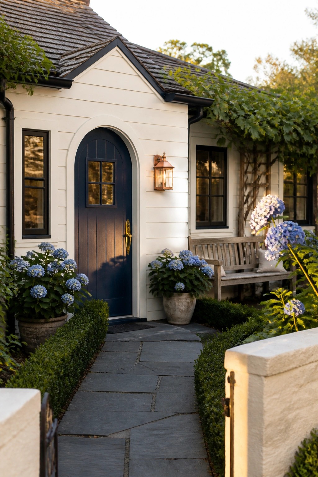 A small white house exterior featuring an arched dark blue front door, black-framed windows, stone pathway, and potted hydrangeas beside a wooden bench.