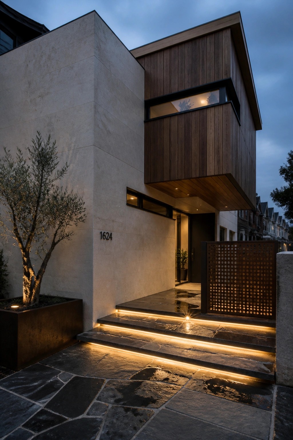A modern house exterior at dusk with light stone walls, dark wood cladding on the upper level, illuminated stone steps, and a small tree in a metal planter.