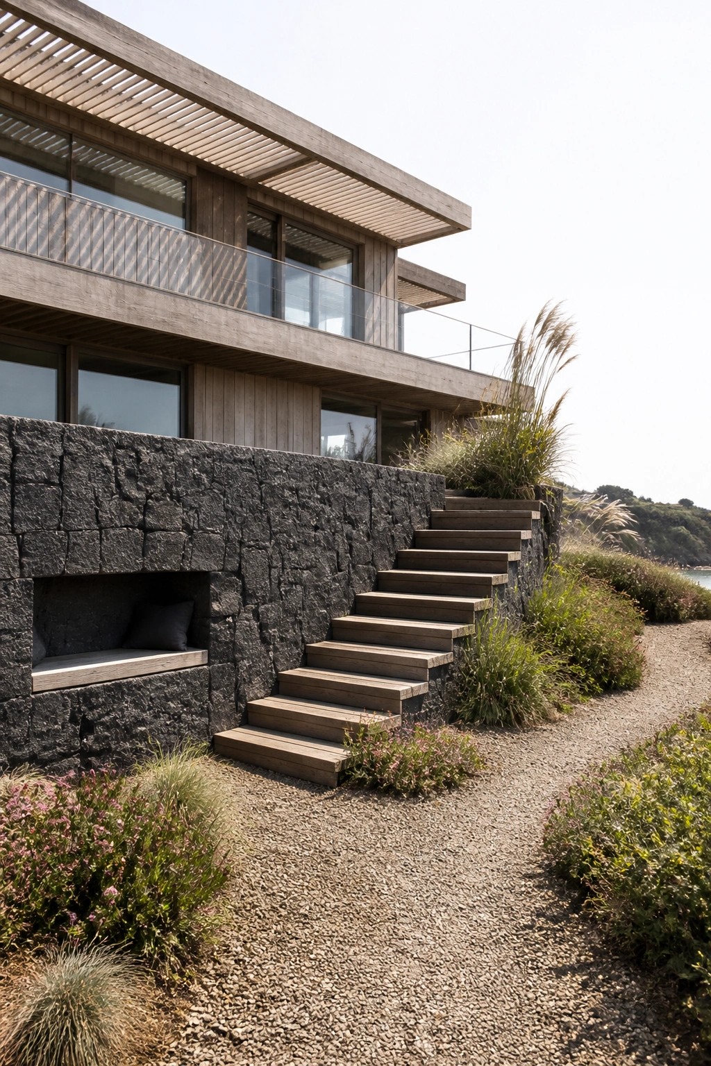 Modern two-story house exterior featuring dark stone walls with a recessed bench, wooden stairs, large windows, and wood cladding against a gravel path and coastal plants.