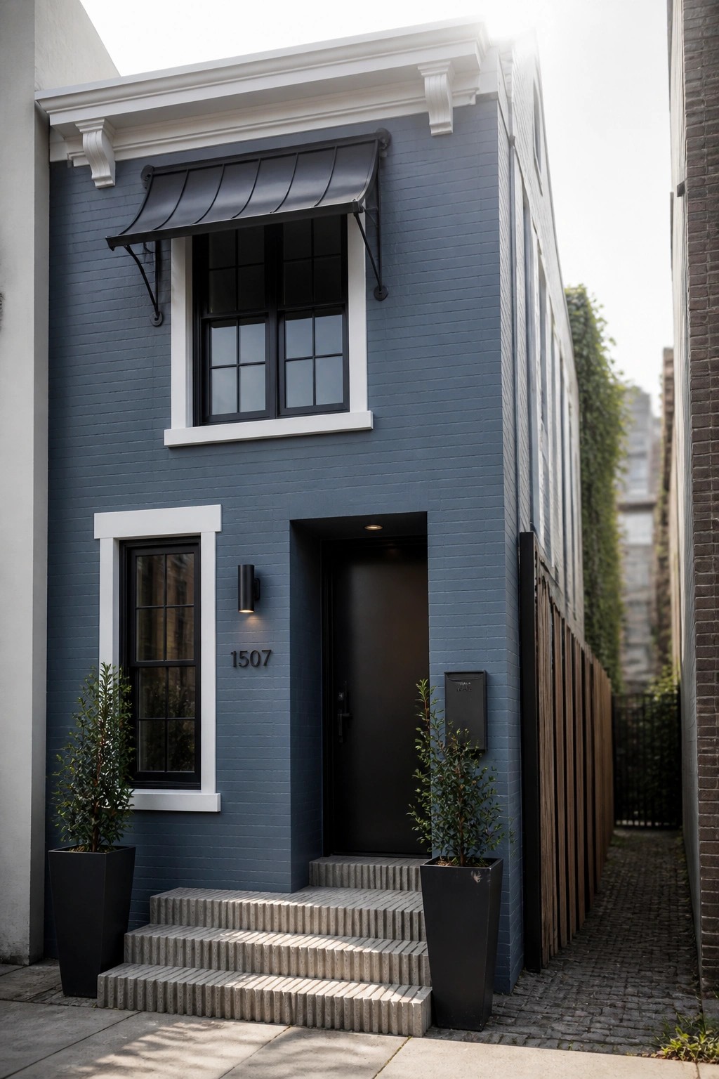 A narrow two-story house with blue-gray painted brick, a black front door, white window trim, and concrete steps flanked by two tall planters.