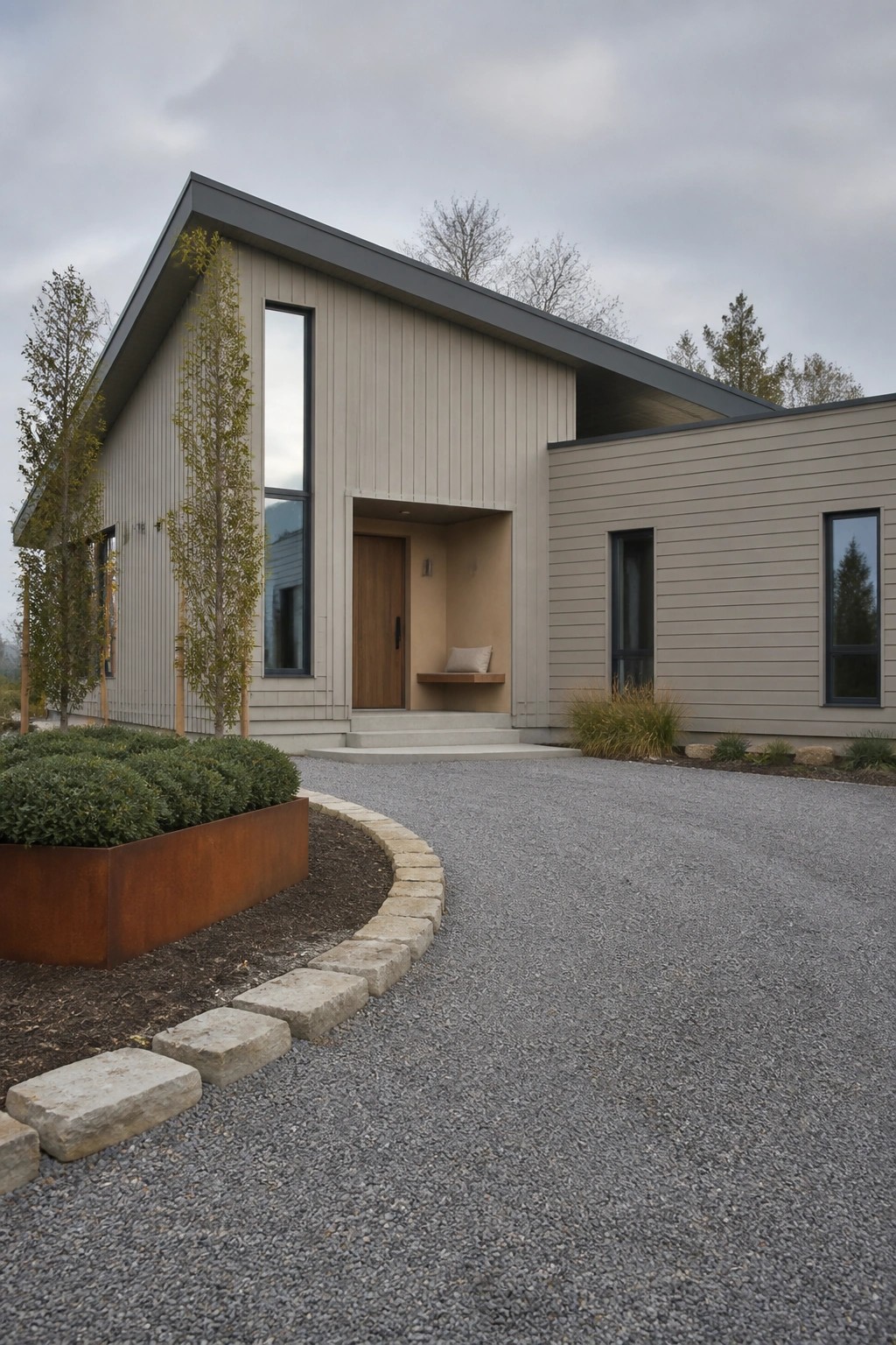 A modern house with light vertical wood siding, a recessed entry containing a wooden door and small bench, tall narrow windows, and a gravel driveway in front.