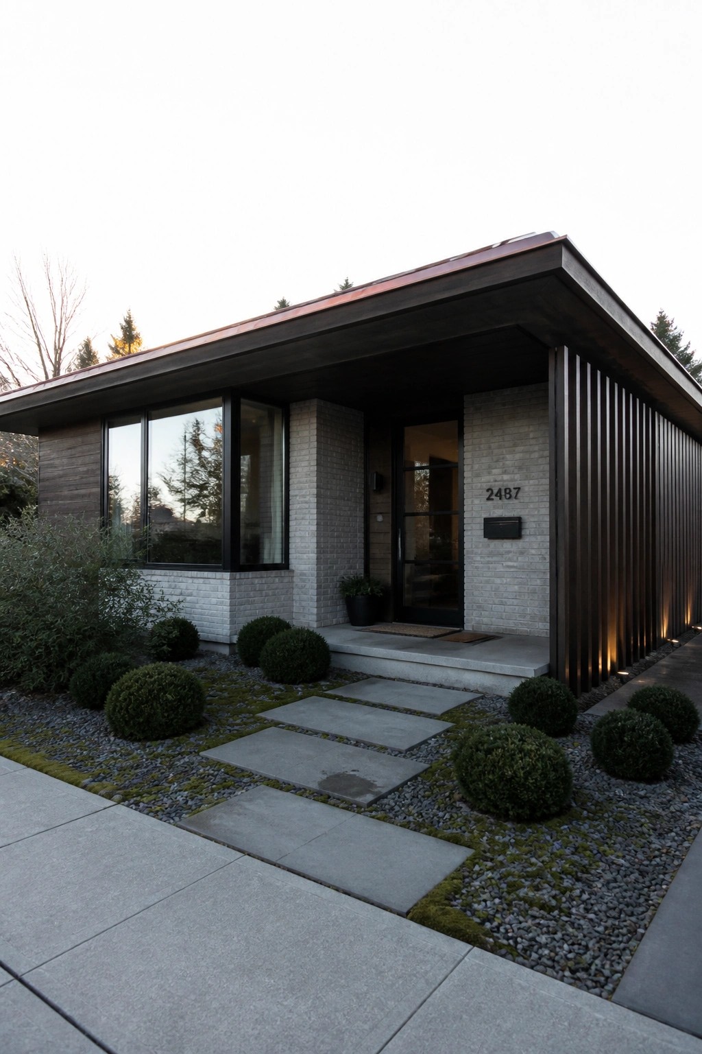 Modern single-story house with a sloped copper roof, gray brick walls, dark wood vertical siding, black-framed windows, and a glass entry door.