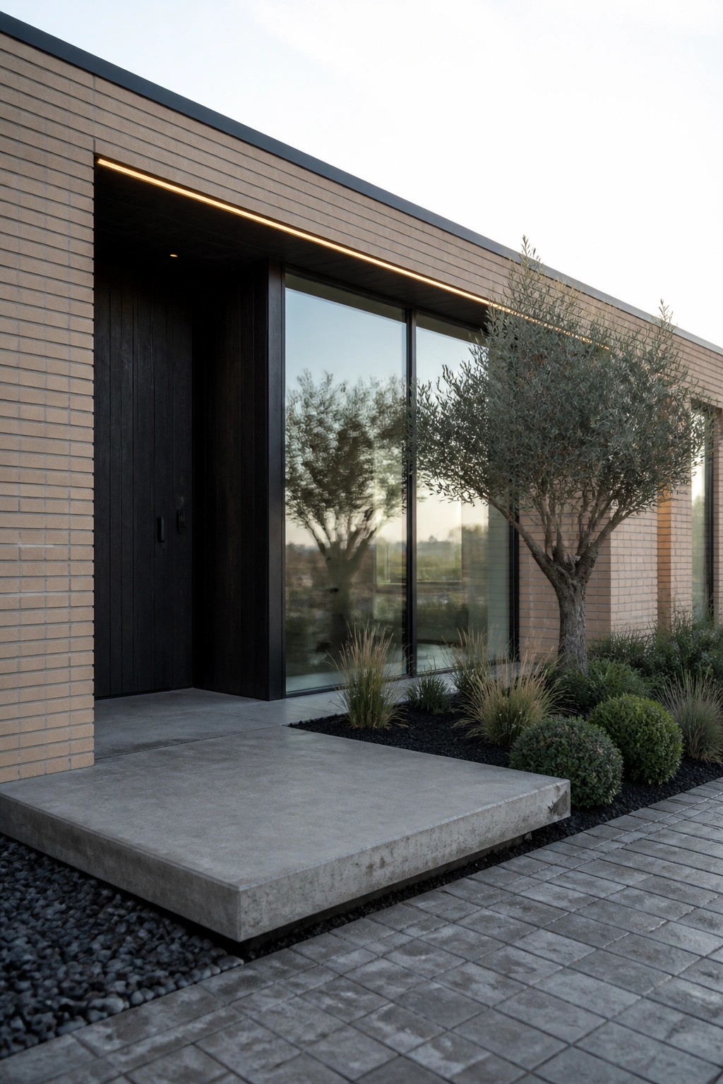 Modern single-story brick house with light tan brick siding, dark wood entry door and panel, large black-framed windows, concrete front step, and small trees in a mulched bed.