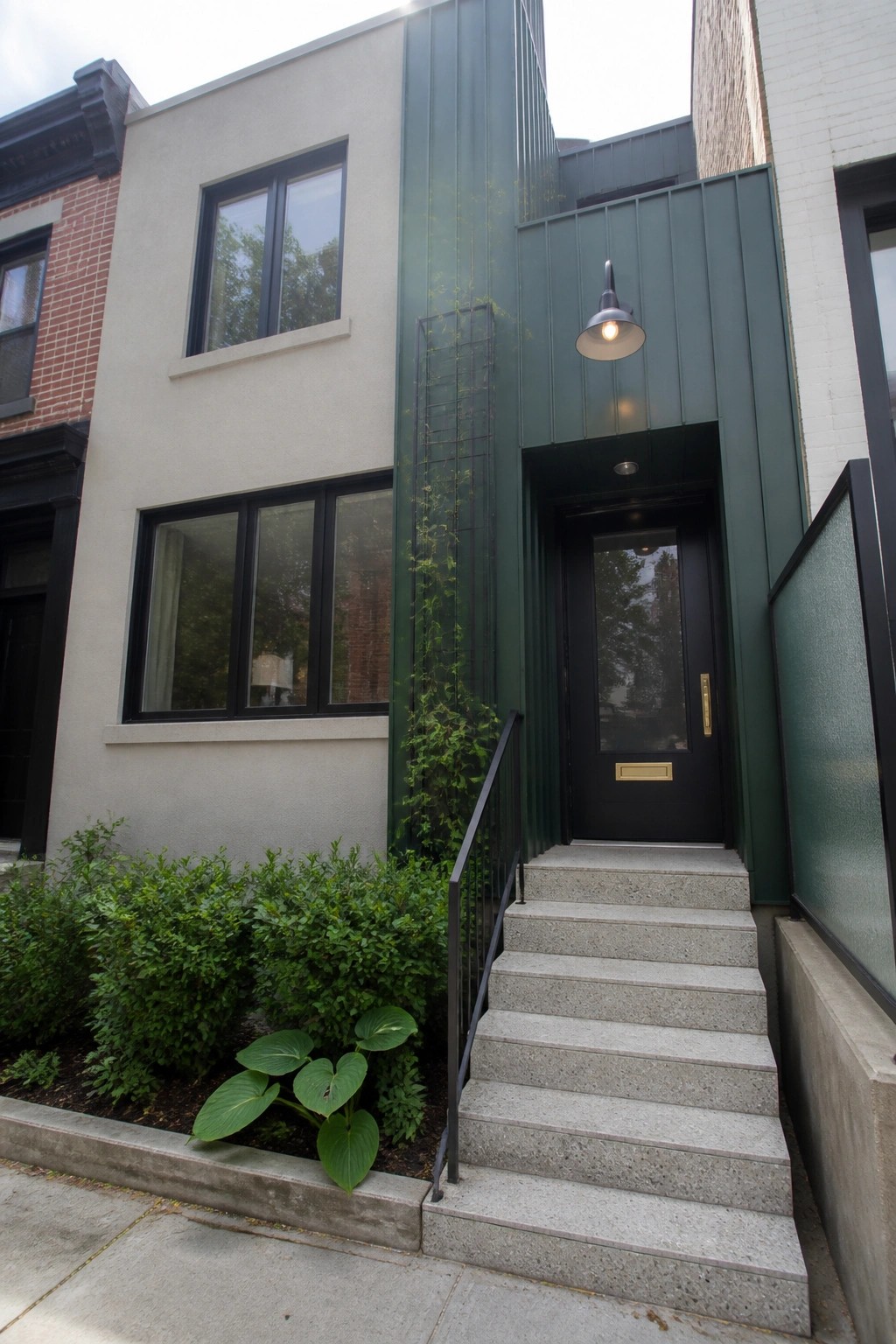 A modern home exterior featuring light beige walls, a vertical dark green metal panel beside the entrance, a black door, concrete steps, and green shrubs in the front bed.
